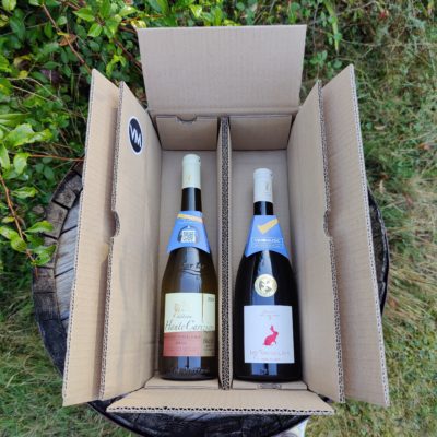 Une bouteille d'Anjou villages avec une bouteille de Muscadet Sèvre et Maine dans une boite en carton posée sur un tonneau.